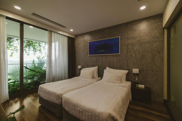 A stylish hotel room featuring twin beds, natural light, and a view of lush greenery.